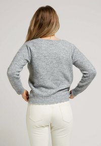 Pull en laine gris avec un col rond et des manches longues ajustées, présentant un ourlet en dentelle et une texture douce. Porté sur un pantalon blanc.