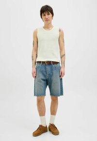 Lichtgekleurde geribbelde tanktop, blauwe denim shorts, bruine riem, bruine schoenen en crèmekleurige sokken. Model staat tegen een effen achtergrond.