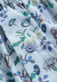 Tissu bleu clair avec un motif floral en tons verts, bleus et violets, présentant un gros plan d'un bouton lavande cousu avec une couture croisée.