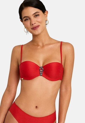 Femme portant un bikini rouge texturé avec des bretelles fines et une attache décorative noire et blanche au centre du haut.