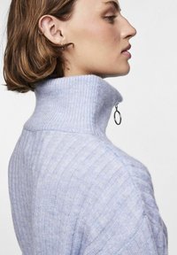 Svetlo modra rebrasta pulover s turtleneck ovratnikom in pol zadrgo ter srebrnim obročkom za zategovanje. Mehka tekstura z izrazitimi šivi.