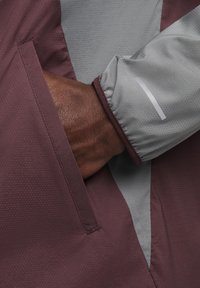 Une main à la peau foncée insérée dans la poche d'une veste perforée bordeaux et grise avec un poignet élastique et une bande réfléchissante sur la manche.