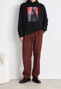 Sudadera negra con estampado gráfico que presenta un rostro, combinada con pantalones marrones de corte holgado y zapatos negros. Texturas suaves, ajuste relajado.