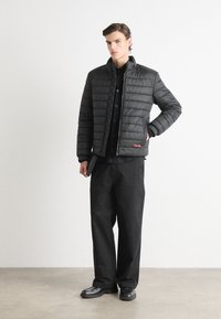 Zwarte pufferjack met horizontaal gestikt ontwerp, hoge kraag en rood merklogo. Gedeeltelijk over een donker shirt gedragen, gecombineerd met zwarte broek.