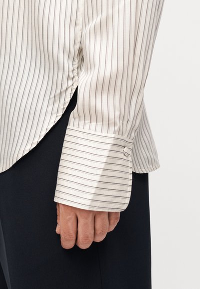 Χέρι με λευκό πουκάμισο μακριάς μανσέτας με λεπτές κάθετες ρίγες, με μανσέτες κουμπωμένες, ακουμπά δίπλα σε σκουρόχρωμο παντελόνι.