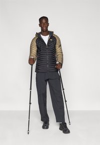 Μαύρο και ταουπέ μονωμένο μπουφάν με φερμουάρ και κουκούλα, σε συνδυασμό με μαύρο παντελόνι και μπαστούνια πεζοπορίας. Ομαλή υφή, καπιτονέ σχέδιο.