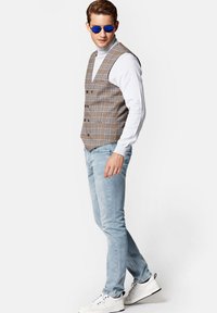 Brązowa kamizelka w kratę na białym golfie, jasnoniebieskie dżinsy i białe trampki, z okrągłymi niebieskimi okularami przeciwsłonecznymi. Widok z boku.