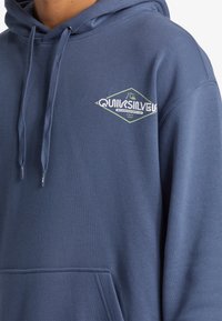 Námornícky modrá mikina s kapucňou vyrobená z bavlneného zmesi. Má predné klokanie vrecko a grafické logo na hrudi.