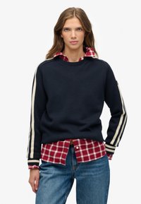 Granatowy sweter z dzianiny z białymi paskami na rękawach, nałożony na czerwoną koszulę w białą kratę, w połączeniu z niebieskimi dżinsami.