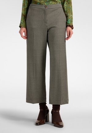 Vrouw draagt een zwart-witte pied-de-poule broek met een hoge taille en wijde pijpen, bruine hakken en donkerbruine sokken, met een groen patroon topje.