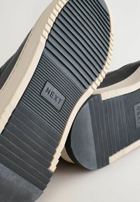 Nærbillede af to sneaker-ydersåler med et struktureret sort gummimønster og en "NEXT"-etiket på bunden, sat mod en lys baggrund.