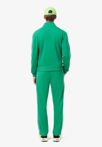 Grøn træningsdragt bestående af en zip-up jakke og bukser. Har ribbede manchetter og sidestriber. Følger med en matchende grøn cap.