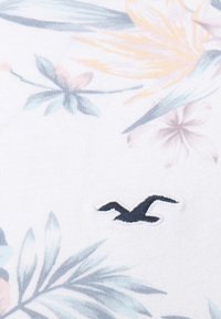 Vitt tyg med ett färgglatt blommönster i nyanser av rosa, blått och gult, med en mörkblå varumärkeslogotyp broderad på det.