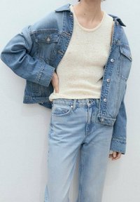 Chaqueta de denim sobre una camiseta de punto color crema, combinada con jeans azul claro. Presenta un corte relajado, detalles de botones y un efecto desgastado.