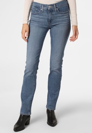 Femme portant un jean en denim bleu taille mi-haute avec un léger effet délavé, associé à des bottines noires et un haut léger à manches longues, debout devant un fond blanc.