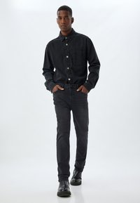 Chaqueta de mezclilla negra con cierre de botones y bolsillo, combinada con jeans negros de corte slim y botas negras hasta el tobillo, de pie contra un fondo claro.