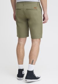 Olivgrüne Shorts mit geradem Schnitt, zwei Gesäßtaschen und einem Leder-Logo-Patch. Kombiniert mit schwarzen High-Top-Sneakern und weißen Socken.