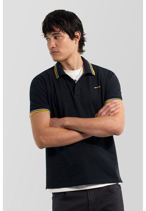 Giovane uomo con capelli scuri che indossa una polo nera con rifiniture gialle, braccia incrociate, guarda di lato su uno sfondo chiaro uniforme.