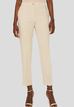Beige skræddersyede ankellange bukser med frontknap og lommer, båret med gyldne højhælede sandaler med strop på en mørk hudfarvet person.