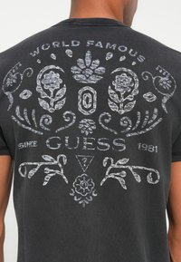 Camiseta negra por detrás con diseños florales y ornamentales en blanco desvanecido, texto "WORLD FAMOUS GUESS SINCE 1981" y un logo de triángulo con un signo de interrogación.
