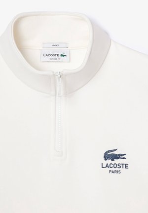 Biała bluza Lacoste z zamkiem do ćwiartki i stójką, z logo krokodyla Lacoste i napisem Paris na lewej piersi.
