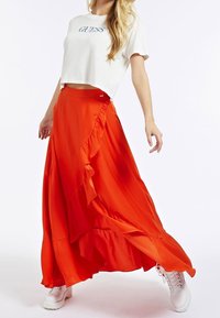Blonde vrouw in witte Guess crop top, met een vloeiende feloranje maxi rok met ruffles en witte veterschoenen, staande op een witte achtergrond.