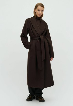 Femme portant un long manteau ceinturé marron foncé, un pantalon noir et des chaussures foncées, debout devant un fond clair avec une main dans la poche.