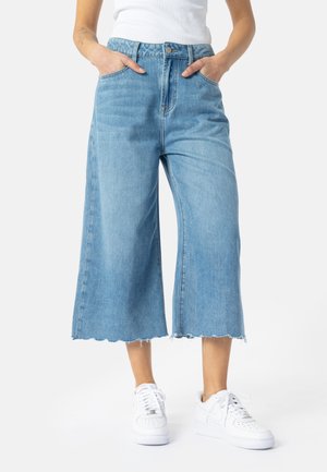 Person iført lyseblå vidde-cropped jeans med frynsete kanter, hvide sneakers og en hvid ærmeløs top, hænderne i lommerne.