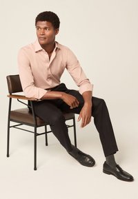 Hellrosa Hemd mit Knöpfen, schwarze Hose und schwarze Loafers mit grauen Socken. Die Person sitzt auf einem modernen Stuhl mit einem Armlehnen aus Holz.