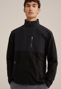 Svart fleecejacka med en solid övre del, hög krage, full dragkedja och två sidofickor med dragkedja. Texturerat tyg och snygg design.