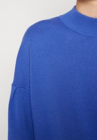 Gestrickter blauer Pullover mit hohem Rippkragen und angesetzten Schulternähten. Glatte Textur mit enger Passform.