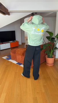 Camisola com capuz verde claro com texto "On Vacation" em branco e gráficos coloridos. Com calças pretas largas. Piso de madeira e plantas interiores visíveis.