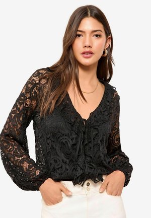 Black lace blouse with long sleeves, featuring a V-neck, ruffle details, and a front tie closure. Textured with intricate floral patterns.