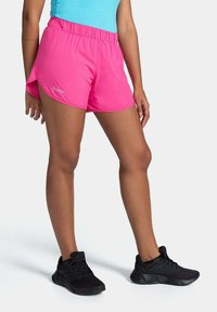 Lyserøde atletikshorts med en bred elastisk talje, sideskår og et lille logo. Sammen med sorte sneakers på en lys baggrund.