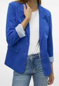 Niebieska marynarka z klapami, zapinana na guziki, z podwiniętymi mankietami w paski, w zestawie z białą bluzką i dżinsami.