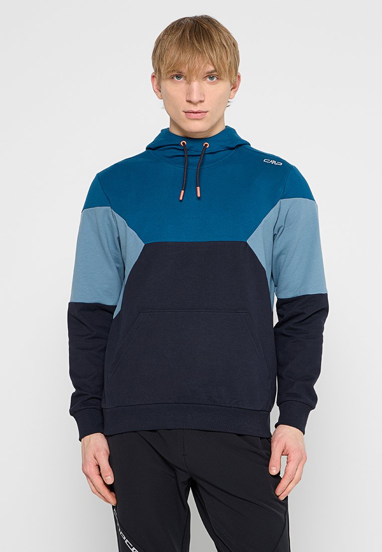 Giovane uomo con capelli biondi lisci, che indossa un hoodie blu e nero a blocchi di colore con una tasca frontale e pantaloni neri, su uno sfondo semplice.