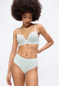 Γυναίκα με αφροχτένισμα που φοράει ανοιχτό μπλε ριμπ ανώριο και εσώρουχα με ψηλή μέση, κοιτάζει προς τα κάτω με το ένα χέρι στη μέση, πάνω σε λευκό φόντο.