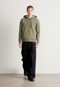 Felpa con cappuccio verde oliva con zip, tasche frontali e coulisse, abbinata a jeans di denim scuro a gamba larga e scarpe nere su uno sfondo neutro.