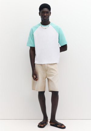 Mand iført en hvid og mintgrøn kortærmet skjorte, beige shorts, en perlehalskæde og sorte flipflops, stående mod en hvid baggrund.
