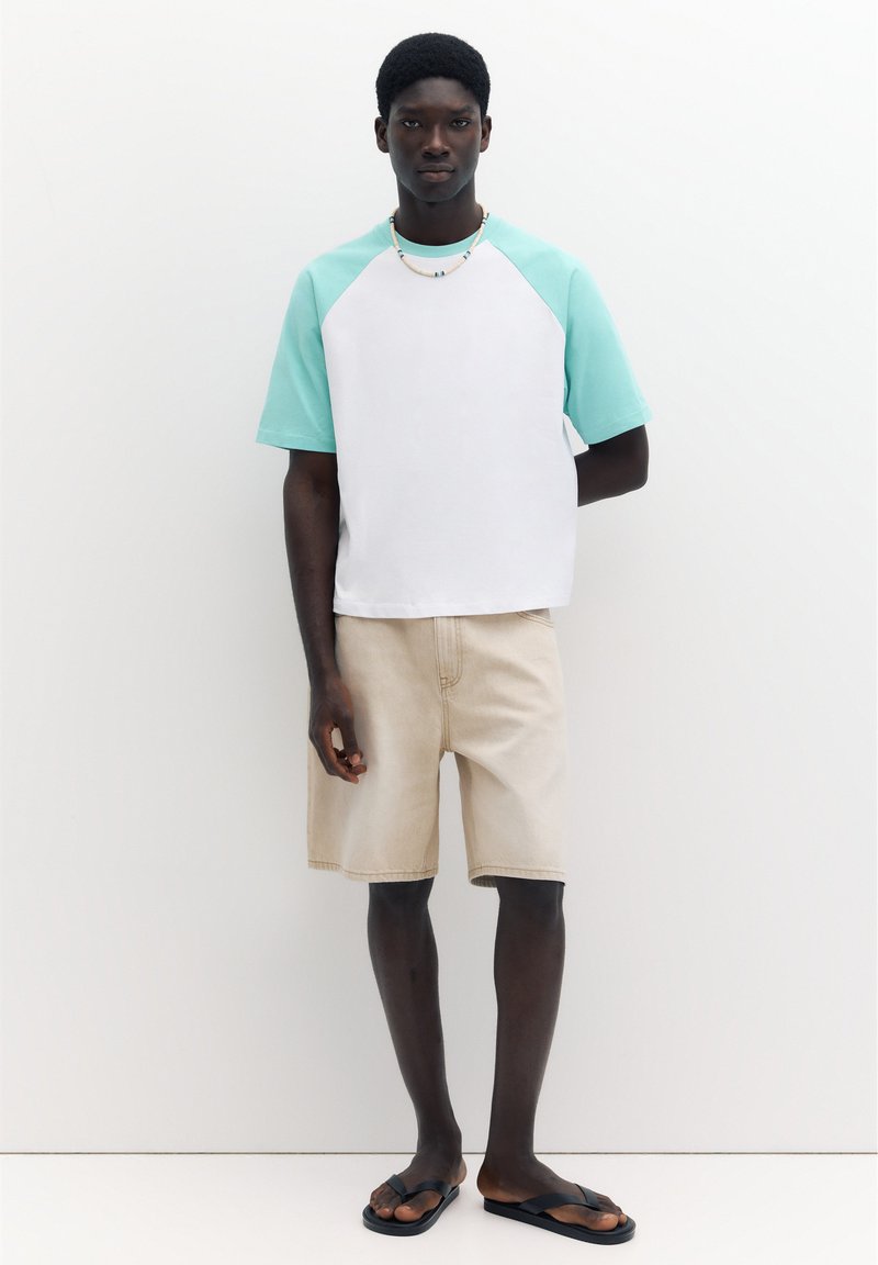 Homme portant une chemise à manches courtes blanche et vert menthe, un short beige, un collier de perles et des tongs noires, debout devant un fond blanc.