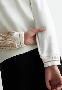 Maglione bianco con maniche in beige lavorato a maglia testurizzata, con accenti dorati e polsino brandizzato che mostra "LIU-JO". Design semplice e informale.