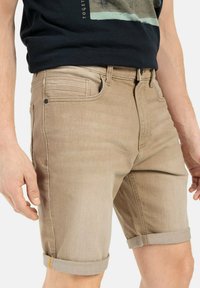 Shorts en denim beige avec une coupe slim, dotés de revers et d'une texture subtile. Fermeture à bouton en métal et poches avant.