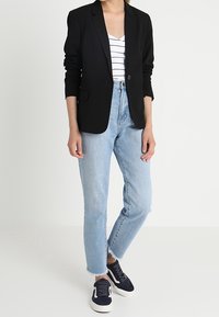Blazer noir à coupe slim, avec revers et un bouton unique, associé à un débardeur blanc à rayures et un jean bleu clair raccourci, complété par des baskets bleu marine.