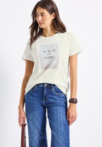 Femme en T-shirt blanc avec le texte "OWN IT", jeans bleus et bracelet marron, tenant un sac à bandoulière marron, regardant sur le côté devant un fond blanc.