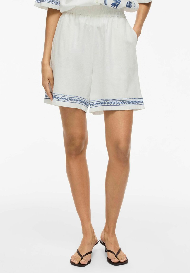 Femme portant un short blanc avec un bord décoratif bleu, une taille élastique, des poches latérales et des sandales noires à talons.