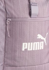 Sac à dos en tissu rayé violet avec une poche zippée à l'avant, des bretelles réglables et un grand logo Puma blanc. Le matériau texturé ajoute de la profondeur.
