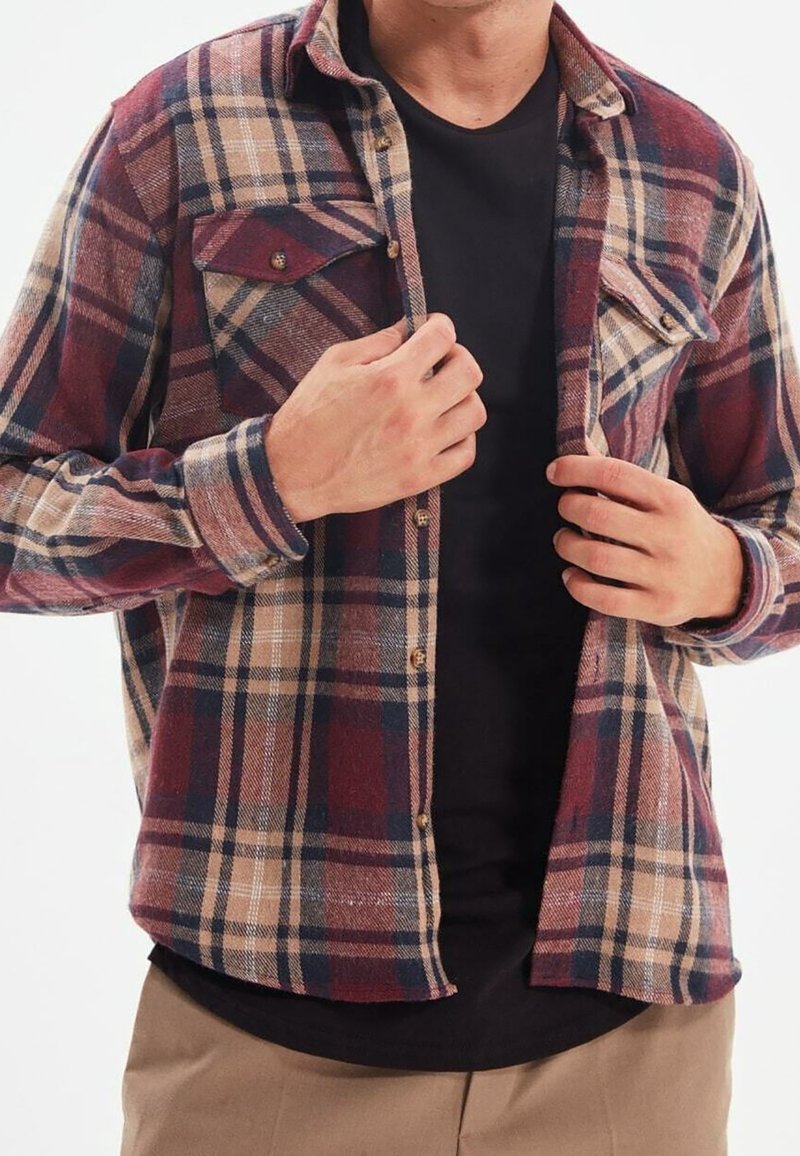 Chemise en flanelle à carreaux rouge et beige, manches longues, poches avant et fermeture à boutons ; portée par-dessus un T-shirt foncé.