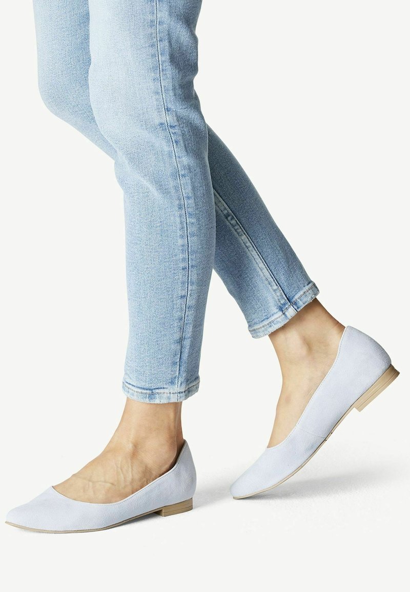 Pieds portant des ballerines pointues bleu clair, assorties à un jean skinny bleu clair retroussé, sur fond blanc.