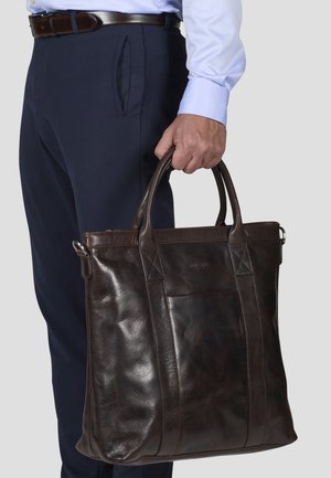Brown leather tote bag with two handles, a front pocket, and smooth texture. Bag held by a person in navy trousers and a light blue shirt.