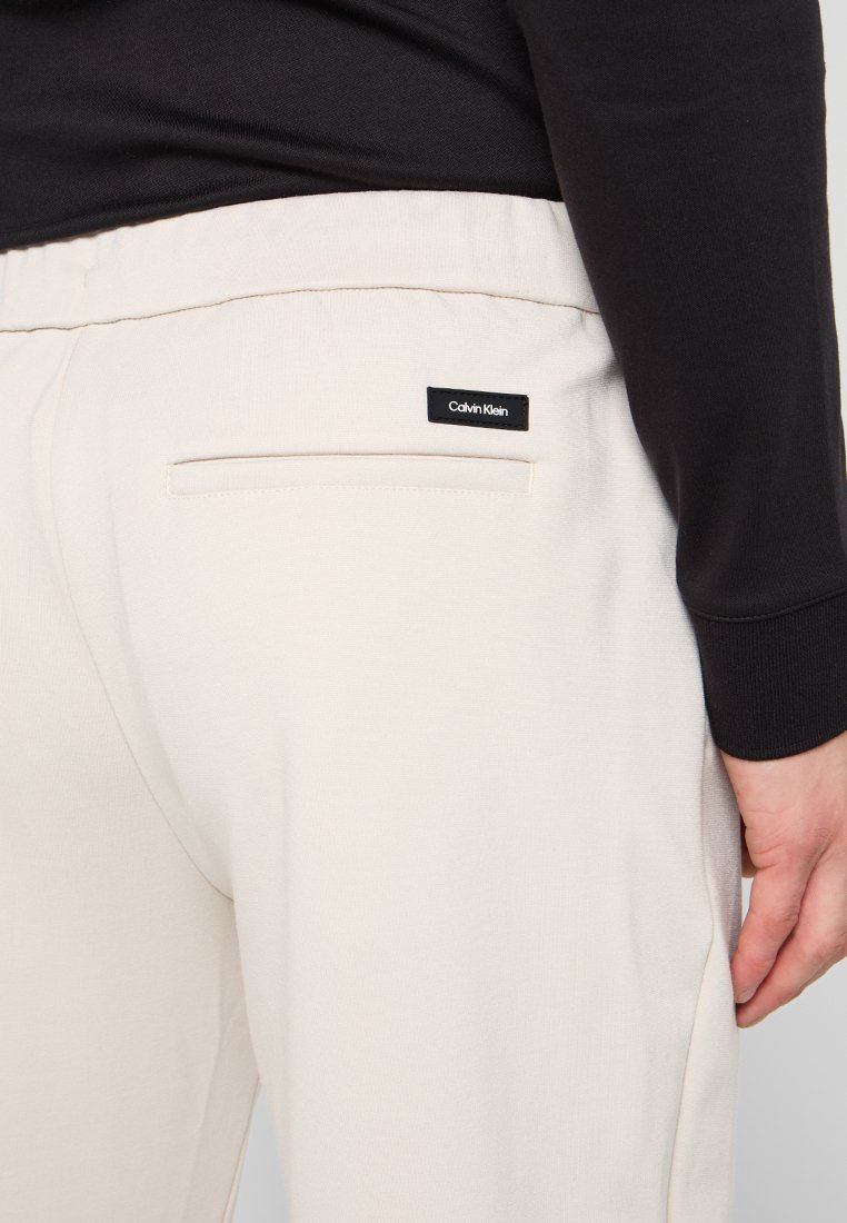 Helle beige Hosen mit strukturiertem Finish, ausgestattet mit einer Gesäßtasche und einem kleinen schwarzen Calvin Klein-Logoetikett in der Nähe des Bundes.